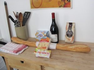 Wooden kitchen table with bottle of wine, baguette, cheese, and other food.