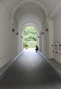 Long outdoor corridor through a building; the walls are white, and green plants are visible through the arched entry at the end. Near the end of the corridor, a person is visible in silhouette sitting on a low curb.