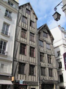 Two tall, narrow half-timbered houses with peaked roofs, windows, and multiple stories. The house on the right is shorter than the house on the left, and the height of the stories varies.