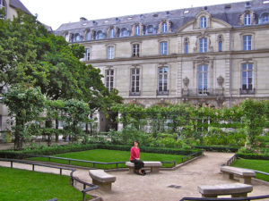 Woman in a red shirt and black skirt sitting on a stone bench surrounded by neat paths and greenery, with a dignified old multi-story stone building filling most of the background