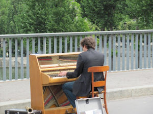Man seated at small piano outdoors, with the railing of a bridge in the background