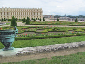 Elaborately designed gardens and small, neatly trimmed evergreen trees, with the palace of Versailles in the background