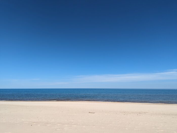 Blue sky with a thin layer of clouds near the horizon. Beneath the sky, Lake Michigan, also blue, and light-colored sand.